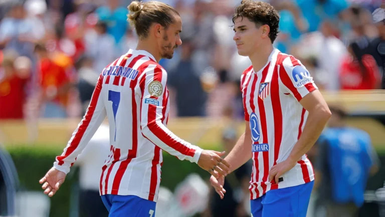 Two Atlético Madrid players in red and white striped kits share a handshake and exchange words during a match.
