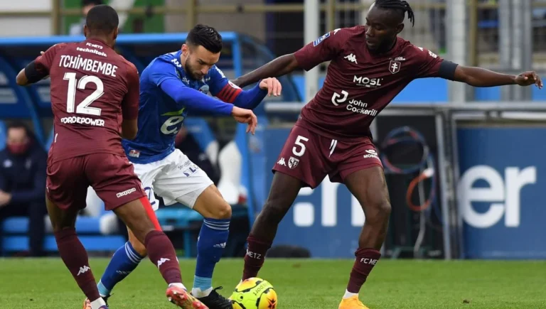 A Strasbourg player in a blue jersey battles for the ball against two Metz players in maroon jerseys during a Ligue 1 match.