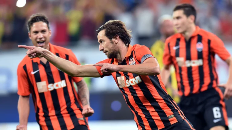 Shakhtar Donetsk player celebrating after scoring a goal, pointing forward, with teammates in orange and black striped kits.