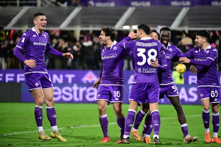 Fiorentina players in purple kits celebrating together on the pitch during a football match.