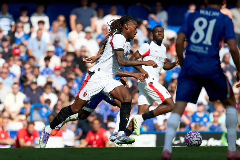 AC Milan player in white drives forward with the ball while Chelsea players in blue defend during a football match.