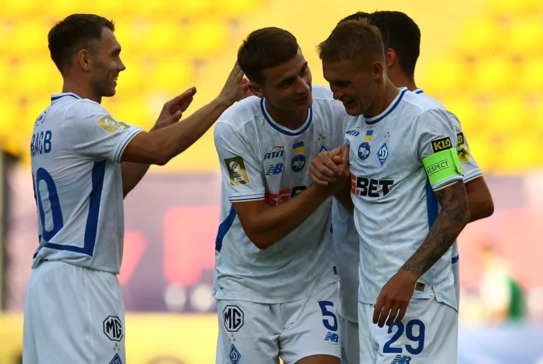 Dynamo Kyiv players celebrate a goal during a match, wearing white kits with blue details and captain’s armband visible.