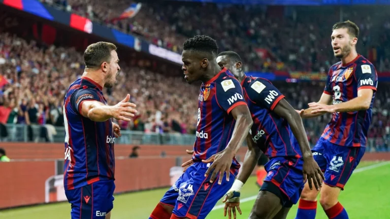 FC Basel players celebrate a goal during a football match, wearing red and blue striped kits, with fans cheering in the background.