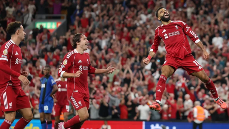 Three Liverpool players in red kits celebrate a goal during a Premier League match. One player jumps into the air with arms raised in joy, while two teammates smile and run towards him. A packed stadium crowd in the background cheers loudly.