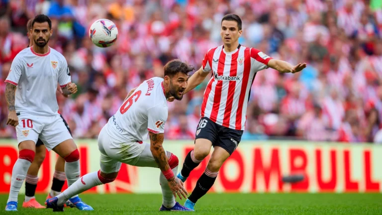 Athletic Bilbao player in red-and-white stripes battles Sevilla players in white during a Spanish La Liga match at San Mamés Stadium.