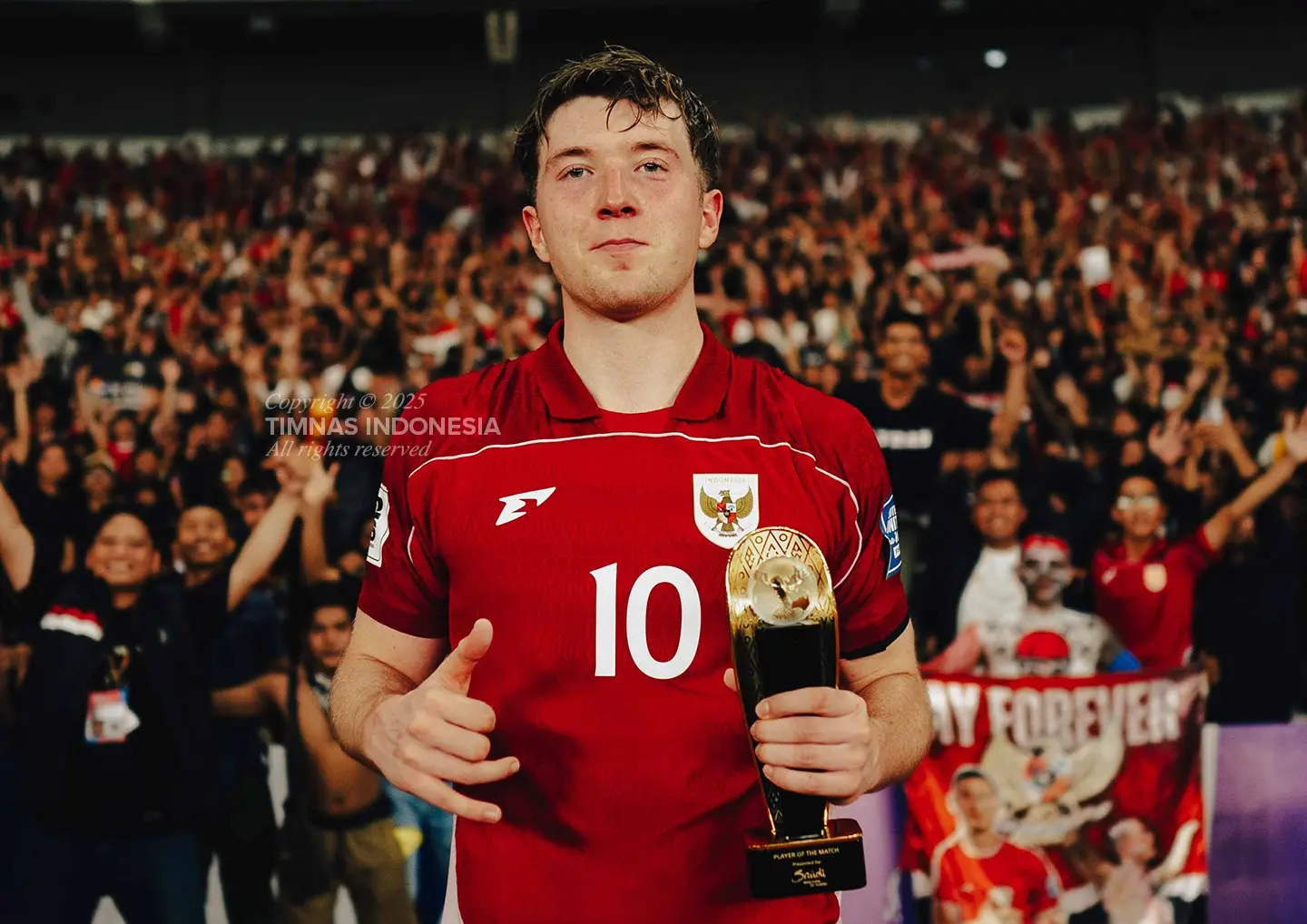 Ole Romeny mengenakan jersey merah sedang memegang trofi penghargaan di lapangan dengan latar belakang suporter yang merayakan.